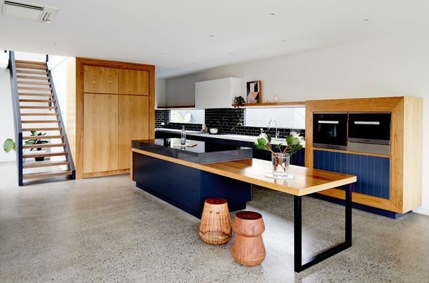 Open-plan contemporary kitchen with a dark navy island, timber breakfast table extension, polished concrete flooring, and a black-and-white tiled splashback. Light timber cabinetry, integrated appliances, and an open stairway contribute to the clean, modern interior.