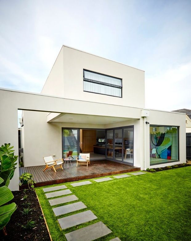 A contemporary two-storey home viewed from its landscaped rear garden. The white rendered facade has a projecting upper volume, broad covered terrace, dark timber decking, extensive glazed doors and windows, and a simple paved path through a manicured lawn. Outdoor seating is arranged on the terrace, with planting along the edges and a colourful artwork visible through the right-hand window.