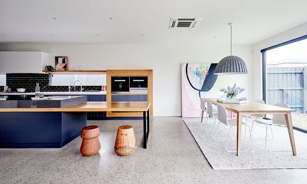Bright open-plan contemporary kitchen and dining area with a dark island, timber benchtops and joinery, integrated appliances, terrazzo-style flooring, and a light dining table beside large glazing. A black-and-white tiled splashback and oversized pendant light provide graphic accents.