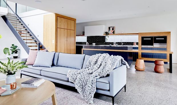Bright open-plan living room with a pale upholstered sofa, textured throw, light timber coffee table, and contemporary kitchen beyond. The space features polished terrazzo-style flooring, white cabinetry, black tiled splashback, timber joinery, and an open timber-and-metal staircase.