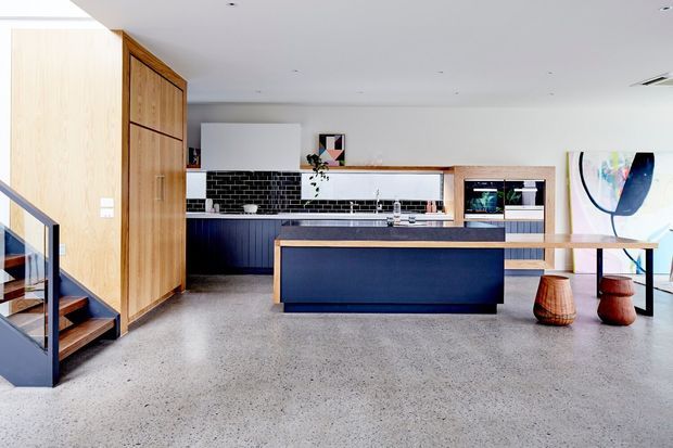 Bright contemporary open-plan kitchen with a large dark island, black tiled splashback, timber cabinetry and pantry doors, integrated appliances, and polished concrete flooring. A timber-and-black stairway is visible at left, with a dining area and artwork at right.