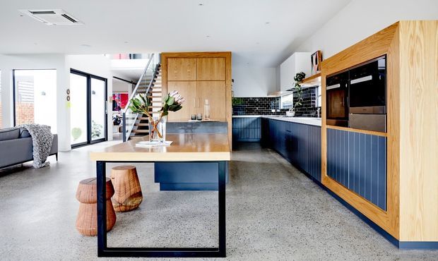 Bright contemporary open-plan kitchen with a large island, integrated timber cabinetry, dark blue fluted lower cabinets, black subway-tile splashback, polished concrete or terrazzo-style flooring, and stairs leading to an upper level. The space includes a small adjacent living area and strong indoor-outdoor glazing.