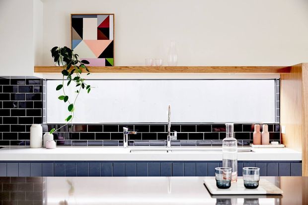 Bright contemporary kitchen with a long white benchtop, black subway-tile splashback, blue-grey tiled cabinetry fronts, timber open shelving and a stainless-steel sink and tap. A geometric abstract artwork, small plants and glassware add restrained decorative accents.