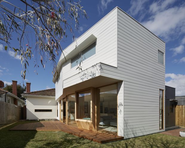 Modern two-storey residential extension with crisp white horizontal cladding, large glazed openings, a projecting upper-level balcony or overhang, and warm timber-framed ground-floor glazing. The house sits beside a timber deck and lawn, with an older tiled-roof dwelling visible behind it.
