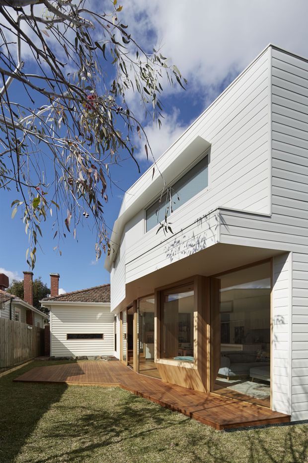 Exterior view of a contemporary two-storey residential extension with white horizontal weatherboard cladding, broad glazed openings, timber-framed doors and windows, and a timber deck wrapping around the ground floor. The building sits in a suburban garden beside an older tiled-roof house, with a projecting upper-level form and clean geometric lines.