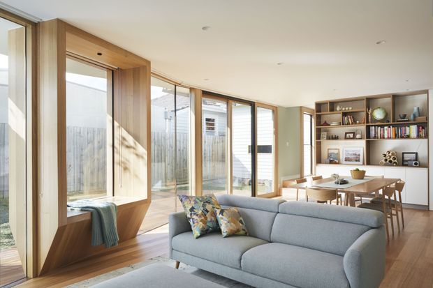 Bright open-plan contemporary living and dining area with a grey sectional sofa, timber-framed window seat, extensive glazing, timber flooring, and integrated timber shelving displaying books and decor. The dining area sits beyond the living space, with a warm neutral palette and abundant natural light.
