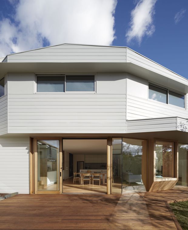 Exterior view of a contemporary two-storey house with white horizontal weatherboard cladding, a low-pitched roof, projecting upper-level form, and extensive timber-framed glazing. Large sliding glass doors open onto a broad timber deck, with a dining area and kitchen visible inside.