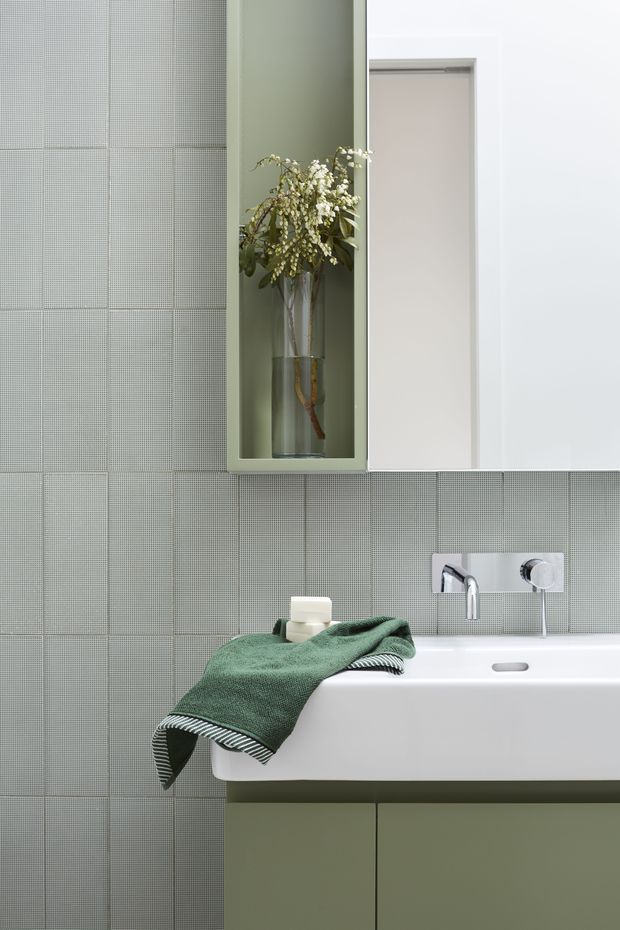 A refined contemporary bathroom vignette featuring a wall-mounted white basin with chrome wall-mounted tapware, an olive-green vanity, and pale green-grey small-format grid tiles. A recessed open shelf displays a tall glass vase with foliage, while a green textured towel adds a muted natural accent.