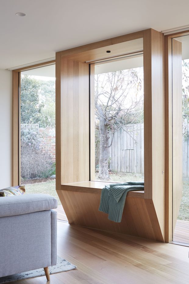 Bright contemporary living room featuring full-height glazing and a distinctive built-in timber window seat with angled cabinetry below. Warm natural wood lines the window reveals and flooring, while a pale upholstered sofa and soft furnishings create a calm, understated interior. The glazing opens toward a private garden and timber deck.