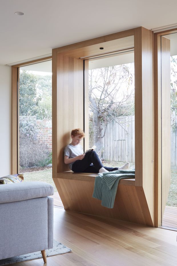 A bright contemporary living room with extensive floor-to-ceiling glazing and a built-in timber window seat. Warm wood joinery frames the windows and forms a sculptural angled base, while a pale upholstered sofa sits in the foreground. A child is reading on the window seat, with a muted green throw draped across it. The room opens visually to a private garden courtyard.