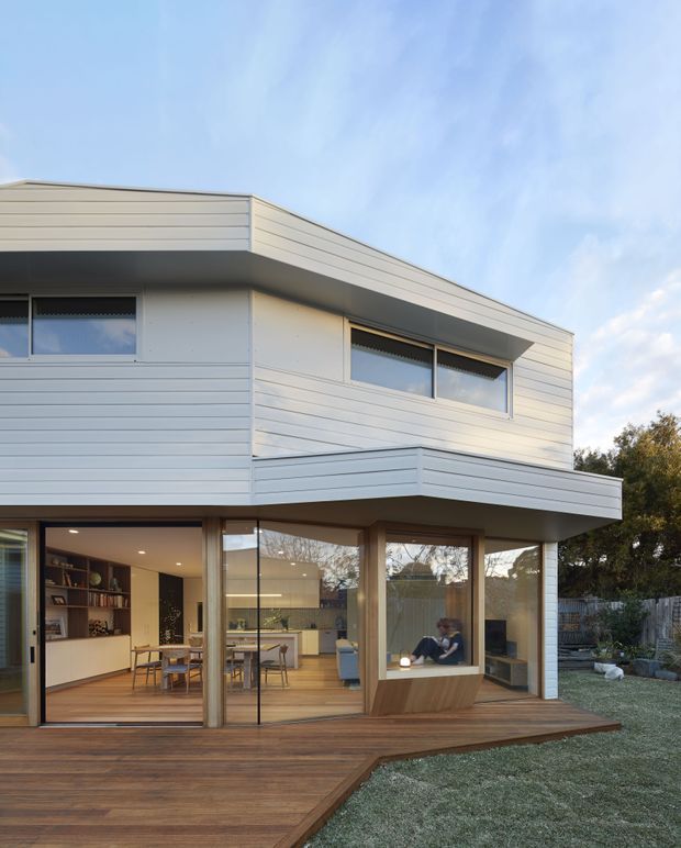 A contemporary two-storey house with a crisp rectangular form, white horizontal weatherboard cladding, broad overhanging roof edges, and extensive ground-floor glazing. The lower level opens onto a large timber deck and landscaped yard, with a visible open-plan kitchen and dining area inside. Upper-level windows have external roller blinds, while the interior appears warm and timber-finished.