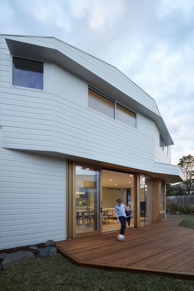 Exterior view of a contemporary two-storey house with white horizontal weatherboard cladding, a steep asymmetrical gable roof, large glazed doors opening onto a broad timber deck, and warm timber-framed openings. The landscaped yard and overcast sky surround the building, while a dining/living area is visible through the glazing.