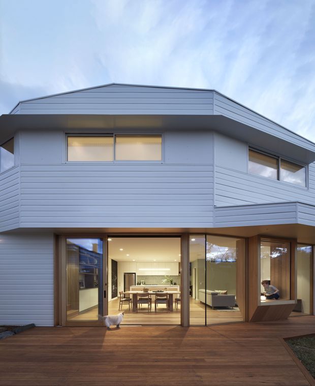 Exterior view of a two-storey contemporary home with pale horizontal cladding, projecting upper level, extensive ground-floor glazing, and a broad timber deck. The open glazing reveals a dining area, kitchen, and living space inside, with a small pet visible near the deck.
