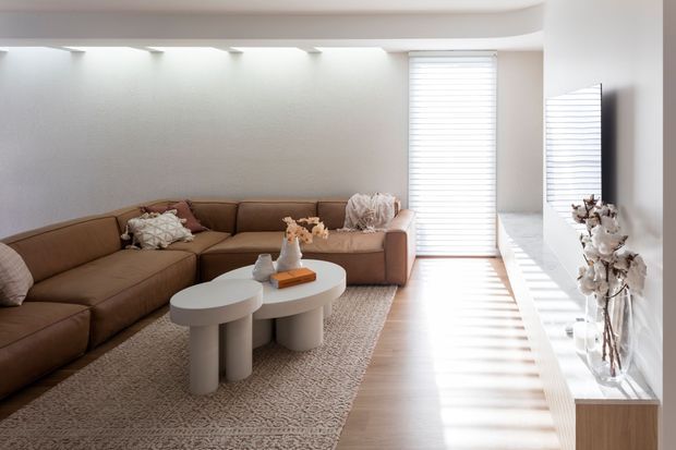 Bright contemporary living room with a large warm brown L-shaped sectional sofa, sculptural light-colored nesting coffee tables, a textured area rug, and a long built-in display ledge beneath a large wall mirror. White walls, recessed linear lighting, and strong daylight through a window with horizontal blinds create a clean, calm interior.