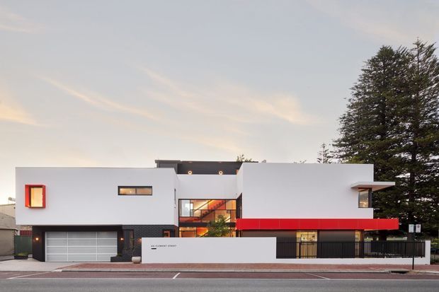 Street-facing photograph of a contemporary multi-level residence with crisp white rectilinear volumes, a bold red horizontal canopy or fascia, dark vertical screening, extensive glazing, and an integrated double garage. Mature trees and neighbouring buildings frame the suburban setting.