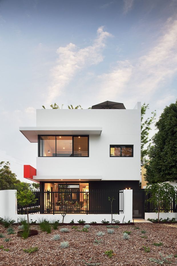 Front exterior view of a compact multi-storey contemporary residence with a crisp white rendered facade, strong horizontal projecting awnings, black-framed glazing, and a black metal front fence. A planted gravel garden occupies the foreground, with warm interior lighting visible through the windows and entry glazing.