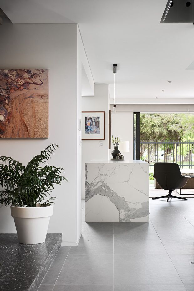 Bright contemporary open-plan interior featuring a freestanding marble-clad kitchen island or counter, large-format grey floor tiles, white walls and ceiling, slim dark window and door frames, and a minimalist lounge or office area beyond. A large potted plant, abstract wall art, and extensive glazing add warmth and visual interest.