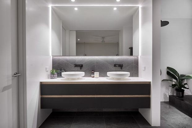 Contemporary double-vanity bathroom with a floating dark wood-toned cabinet, white vessel basins, pale stone benchtop, grey tiled splashback and floor, and a large illuminated mirror. A partial shower area and indoor plant are visible at the right.
