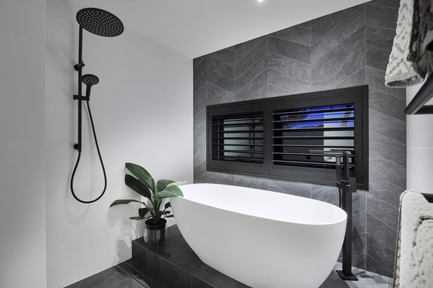 Contemporary bathroom featuring a freestanding white bathtub, matte black shower and tapware, dark grey tiled feature wall, black-framed louvred window, and a potted plant on a dark tiled plinth.