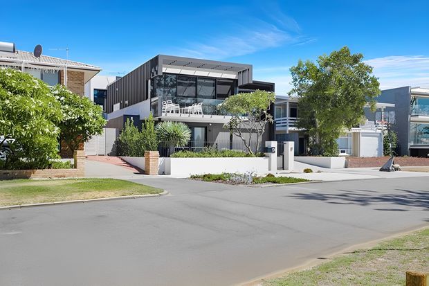 Street-level exterior view of a contemporary multi-storey residence in a suburban neighbourhood. The house has a strong rectangular form, dark vertical cladding, extensive glazing, a projecting upper-floor balcony with white outdoor furniture, pale rendered walls, and integrated front landscaping. Adjacent modern homes and the residential street are visible.