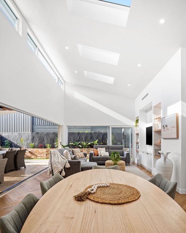 Bright, open-plan living and dining area with a large light-wood dining table in the foreground, upholstered dining chairs, a grey sectional sofa, and built-in white media shelving. The room has a very high sloping ceiling with multiple skylights and recessed downlights, extensive glazing, and a connected outdoor entertaining area visible beyond the sliding doors.