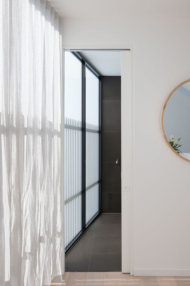 Bright, minimalist interior view toward a compact bathroom or shower area through a white-framed doorway. The space features dark large-format tiled walls and floor, black-framed glazing or a screened partition, and strong linear sunlight shadows. Sheer white curtains and a round timber-framed wall mirror are visible in the foreground.