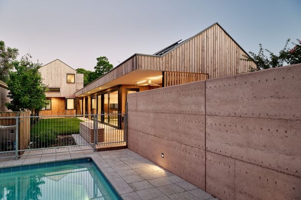 Exterior view of a contemporary residential property at dusk, featuring steep gabled forms clad in vertical timber boards, extensive glazing, a projecting covered walkway, and a swimming pool enclosed by a paved terrace and concrete privacy wall. The composition highlights the building facade, courtyard-like outdoor space, and pool.