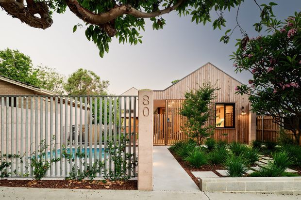 Front exterior view of a contemporary single-storey residence with a timber-clad gabled facade, vertical slatted entry screen, pale rendered entry pillar, low masonry garden wall, landscaped planting, and a partially screened swimming pool behind a vertical metal fence.