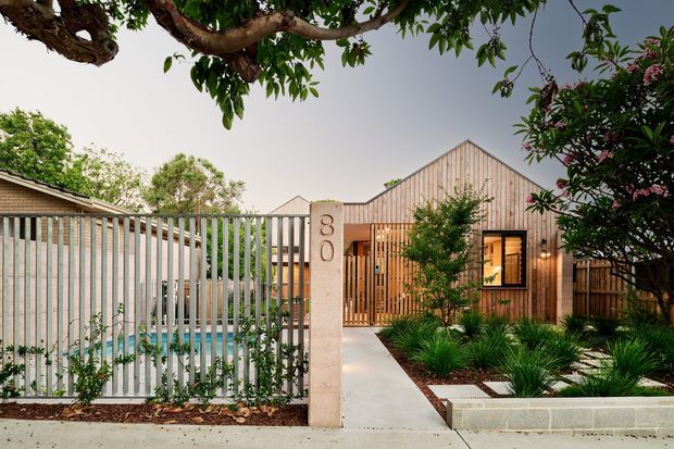 Street-facing view of a contemporary single-storey residence with a pale vertical timber-clad gabled facade, screened entry gate, concrete walkway, landscaped garden beds, and a partially visible swimming pool behind a vertical metal fence. Mature trees frame the property.