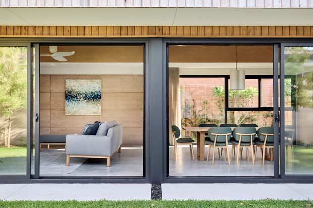 Rear exterior view of a contemporary single-storey home with full-height dark-framed glazing opening onto a living room and dining area. The facade combines pale vertical cladding, timber-lined soffits, and large sliding glass doors. Inside, the living room has timber wall panelling, a sofa, rug and artwork, while the adjacent dining area features a timber table, upholstered chairs, pendant lighting and expansive windows.