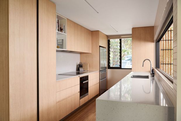 A compact, modern galley kitchen with extensive light timber cabinetry, integrated shelving, a white speckled stone island or peninsula benchtop, stainless-steel sink and tap, integrated appliances, and a dark timber floor. Large windows and louvred glazing provide natural light and views to greenery.