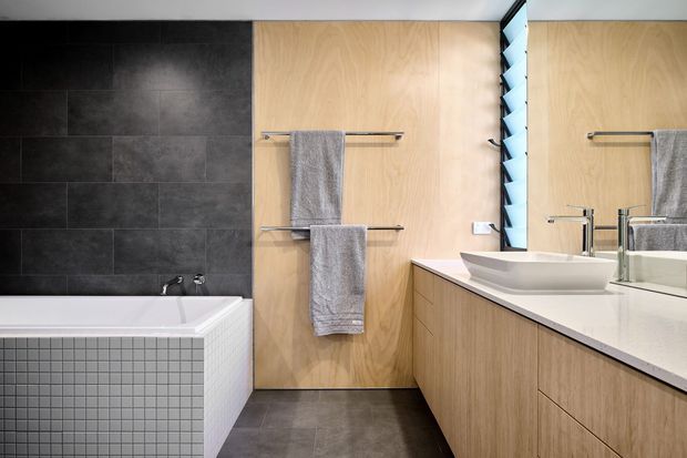 Contemporary bathroom with a freestanding-style white bathtub, dark charcoal wall tiles, pale timber wall panelling, grey floor tiles, and a long timber vanity with a white benchtop and vessel basin. A louvred window beside a large mirror brings in natural light, while simple chrome towel rails and tapware create a clean, restrained fit-out.