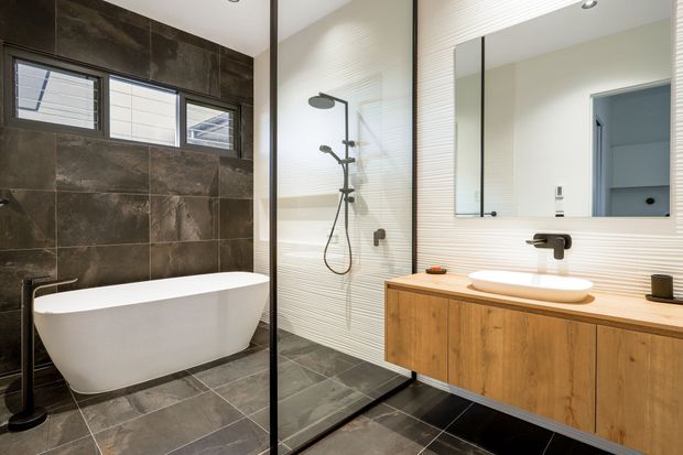 Contemporary bathroom with a freestanding white bathtub, walk-in glass shower, wall-mounted timber vanity with vessel basin, large mirror, dark stone-look wall and floor tiles, and light textured wall tiling. Black fixtures and window frames provide contrast.
