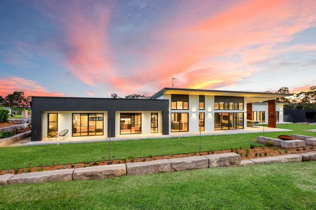 A single-storey contemporary residence photographed from the landscaped front yard at sunset. The building has a low, flat roofline, dark charcoal cladding, white rendered walls, extensive floor-to-ceiling glazing, and a covered entrance or outdoor living area. Terraced stone retaining walls and manicured lawn frame the foreground.