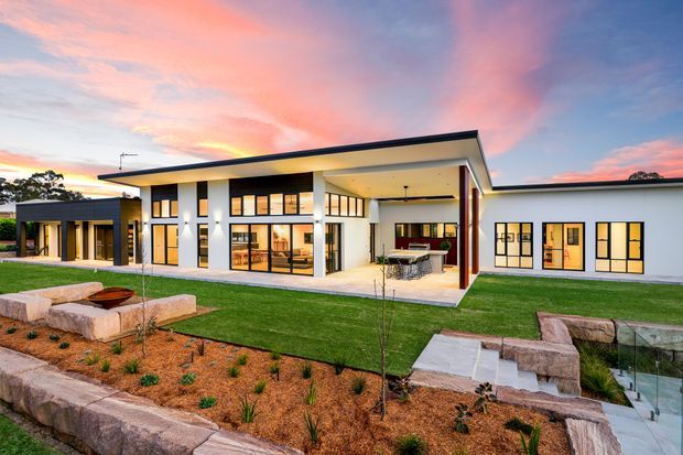 Wide exterior view of a contemporary single-storey residence at dusk, featuring white rendered walls, dark metal rooflines, extensive black-framed glazing, a covered outdoor entertaining area, manicured lawn, and landscaped sandstone garden terraces. Warm interior lighting creates an inviting glow beneath a dramatic pink and blue sunset sky.