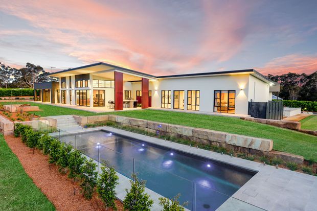 Twilight exterior view of a low, contemporary single-storey residence with a broad flat roof, extensive floor-to-ceiling glazing, white rendered walls, and warm interior lighting. The landscaped property includes a rectangular illuminated swimming pool, paved pool surround, raised stone retaining walls, lawn, and clipped planting.