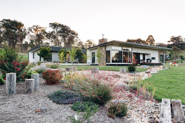 Single-storey contemporary residential home set within a landscaped garden, featuring a low-pitched roof, broad glazed openings, pale rendered walls, dark window frames, and a prominent stone-clad chimney or feature wall. The foreground contains layered ornamental planting, gravel and pebble garden beds, timber edging, and lawn, with mature trees in the background.