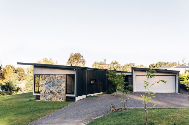 Single-storey contemporary residential exterior with a low, flat-roofed composition. The dark charcoal-clad central volume connects a smaller pavilion featuring a prominent irregular natural-stone wall and extensive glazing to a detached-looking garage volume with a wide white sectional door. A gravel driveway, lawn, young trees, and established landscaping surround the house.