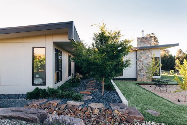 Exterior view of a contemporary single-storey residence with clean white wall panels, dark metal roof edging, extensive glazing, and a prominent stone-clad chimney volume. The landscaped front garden includes gravel beds, native-style planting, a stepping-stone path, layered natural rockwork, and a small outdoor seating area.