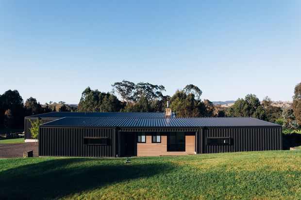 A low, single-storey contemporary rural dwelling with a long rectangular form, dark vertically ribbed metal-clad walls, and a shallow corrugated metal roof. The central entrance and warm timber-clad section contrast with dark window openings, while a chimney projects above the roof. The house sits in an expansive grassy setting with mature trees and distant rolling hills.