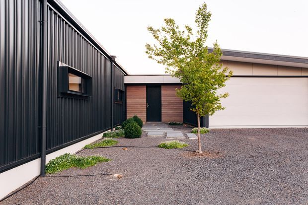 Modern residential exterior courtyard showing a dark vertically ribbed metal-clad building at left, a timber-clad entry volume with a dark door in the centre, and a pale garage structure at right. The landscaped forecourt uses gravel, concrete stepping slabs, low shrubs and a small deciduous tree.