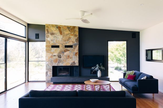 Bright, spacious contemporary living room with floor-to-ceiling glazing, a dark feature wall, and a prominent irregular natural-stone fireplace surround. Two dark blue sofas face a low timber coffee table and television, with a patterned rug adding colour. Timber flooring, white ceiling, ceiling fan, and a view to the outdoors complete the room.