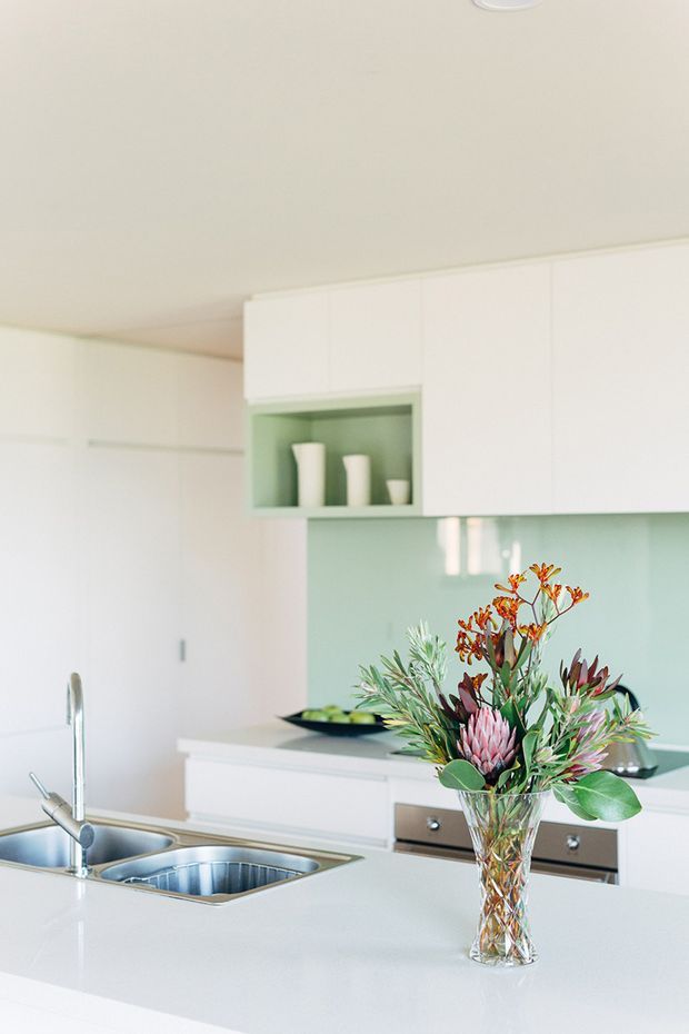 Bright, airy contemporary kitchen with white flat-panel cabinetry, a pale mint-green backsplash and open display niche, stainless-steel double sink and tap, and a white island benchtop. A glass vase of flowers sits on the island in the foreground.