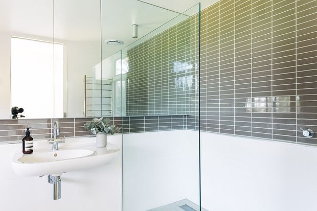Bright contemporary bathroom with a wall-mounted white basin and chrome tapware, a frameless glass shower enclosure, a white bathtub, and grey-green horizontal rectangular wall tiles with white grout. A large mirror and simple white surfaces create an airy, clean appearance.