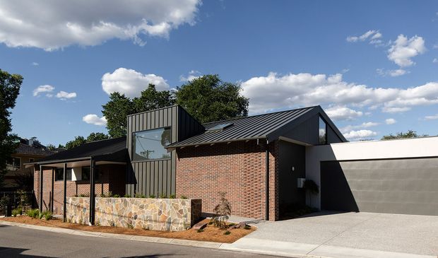 Street-facing view of a contemporary single-storey residence with a mix of dark vertical metal cladding, exposed red brick walls, a steeply pitched dark metal roof, and a broad integrated garage. A low irregular natural-stone wall defines the front garden and covered entry, while large glazing and strong geometric roof forms give the facade a modern architectural character.