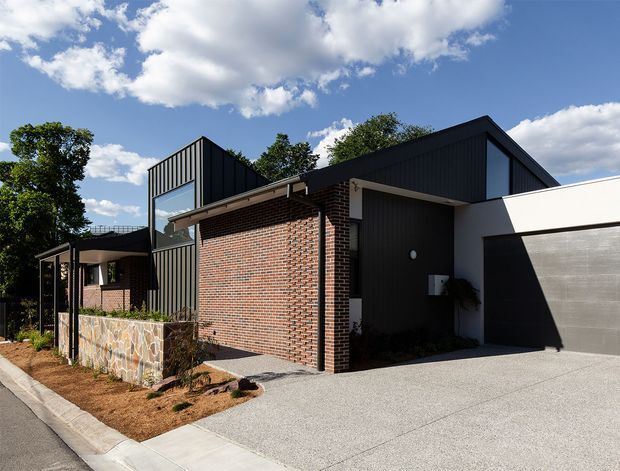 Street-facing view of a contemporary multi-level residence with a combination of dark vertical metal cladding, exposed red brickwork, extensive glazing, and a prominent pitched roof form. The home includes an integrated double garage, concrete driveway, landscaped front garden, and a low decorative stone wall. Bright sunny conditions and mature trees surround the property.