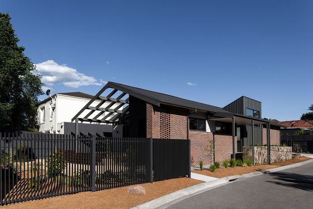 Street-facing view of a contemporary single-storey residence with a dark metal-clad and red-brick facade. The home features a low-pitched roof, large glazed openings, a projecting covered entry or outdoor structure with exposed steel framing, a tall dark metal privacy fence, and a low stone retaining wall. The setting is an established suburban streetscape under clear skies.