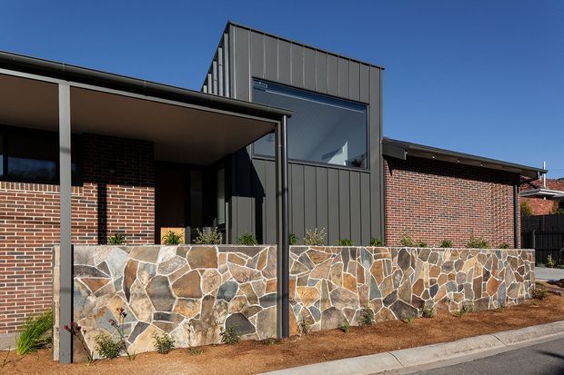 Exterior view of a low-rise brick residence with a prominent contemporary dark metal-clad vertical extension and large glazed window. A broad flat-roofed covered entry or veranda projects across the front, while an irregular natural-stone retaining wall and planted strip run along the street edge.