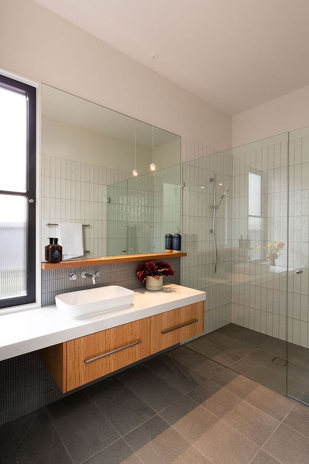 Contemporary bathroom with a walk-in glass shower, wall-mounted timber vanity, white vessel basin, large mirror, pale vertical wall tiles, dark floor tiles, and a black-framed window. Minimal pendant lighting and warm timber joinery add contrast to the neutral palette.