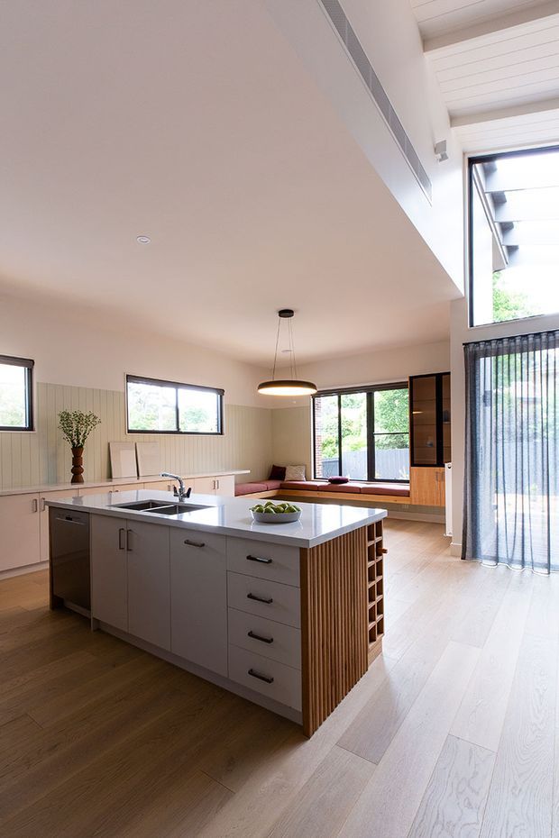 Bright, open-plan contemporary kitchen with a large central island, integrated sink and dishwasher, white benchtop, pale cabinetry, and a timber slatted wine-storage end panel. The kitchen opens to a casual living area with built-in window seating and large glazed doors, while a high white-lined ceiling and abundant natural light create an airy feel.